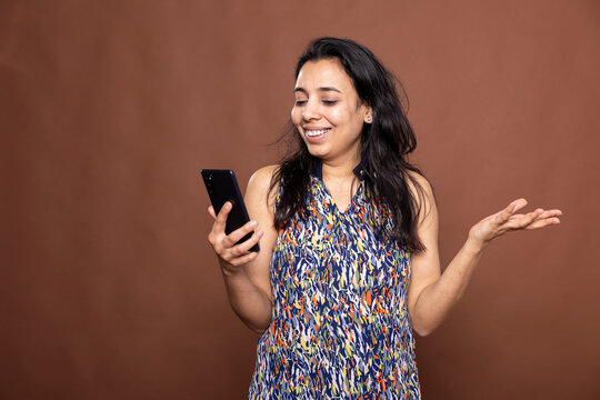 Joyful woman enjoying phone conversation, smiling and laughing while looking at smartphone screen. Gorgeous indian lady in casual clothing reading pleasant text message or watching funny video.