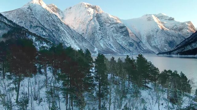Aerial footage capturing the majestic alpine mountain range of Goksoyra in Eresfjord, Norway, bathed in the warm glow of golden hour. The scene features snow covered peaks, a serene fjord, and lush fo