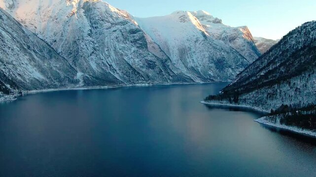 An aerial view captures the serene beauty of a snow covered fjord in Norway during the golden hour of winter. Majestic, snow capped mountains rise dramatically around the tranquil waters, reflecting t