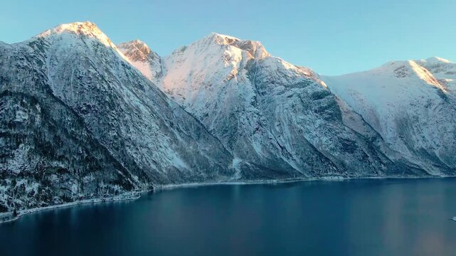 Aerial footage capturing the serene beauty of Eresfjord, Norway, during the golden hour of winter. Snow covered mountains reflect in the calm, deep blue waters of the fjord, bathed in warm, soft sunli