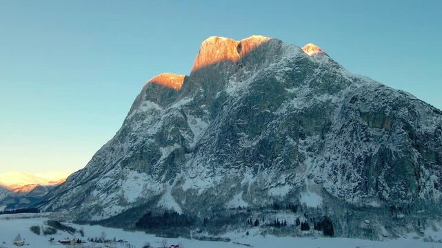 aerial footage captures the stunning winter landscape of Eresfjord or Eikesdalen in Norway, showcasing a snow covered mountain bathed in the warm, golden light of sunrise or sunset.