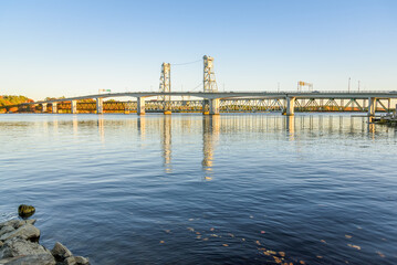 Modern concrete highway bridge running parallel to a railway lift bridge over a large river at sunset in autumn. Reflection in water. Bath, ME, USA.