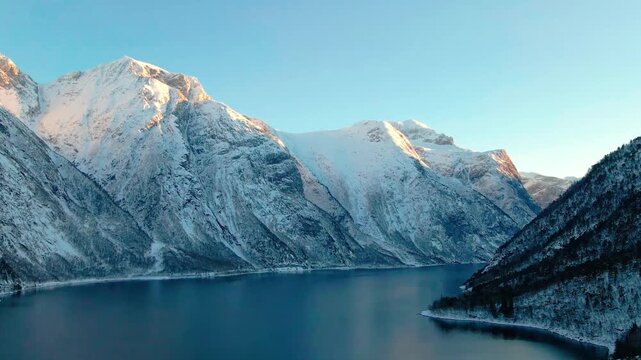 aerial or drone shot captures the serene beauty of a Norwegian fjord during winter's golden hour. Snow covered mountains rise dramatically from the calm, reflective waters, illuminated by the soft, wa