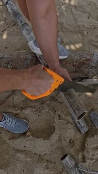 Vertical video. Hands Using Handsaw to Cut Wooden Log on Sandy Beach, Close Up from Overhead Perspective, Outdoor Crafting Concept