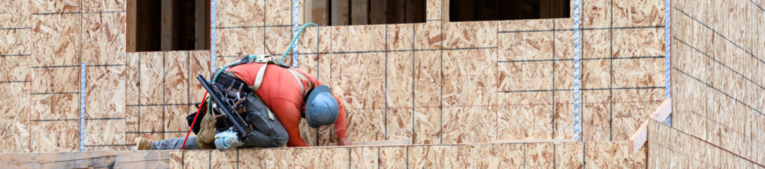 Construction worker in safety equipment using a pneumatic nail gun on the exterior wall of a new...