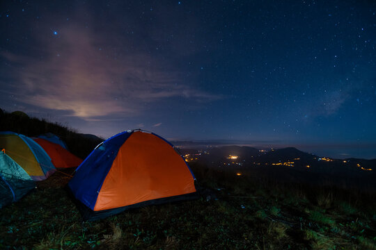Orange and blue camping tent on a green field with mountain silhouettes in the background during a blue starry night