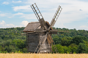 An old wooden windmill on a hill, surrounded by tall grasses and rural vegetation Distant rolling...