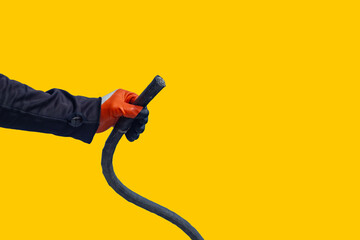 A worker in an orange jacket and gloves holds a black electric cable against a yellow background.