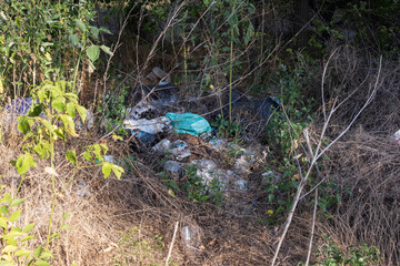Pile of garbage in nature, including plastic bottles, bags, broken glass, and construction debris among dry grass and trees. Environmental pollution, waste management problem.