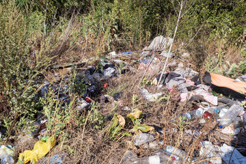 Pile of garbage in nature, including plastic bottles, bags, broken glass, and construction debris among dry grass and trees. Environmental pollution, waste management problem.