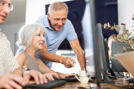 Interested senior woman attending computer class, learning basics of digital technology under guidance of positive aged male teacher standing nearby