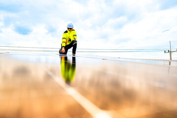 A technician in safety gear inspects solar panels on a metal rooftop, holding a tool. The scene...