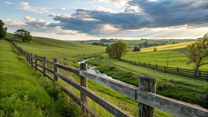 Fototapeta premium Weathered wooden fence stretching along lush green fields on rolling hill countryside farm, under cloudy blue sky with amazing sunlit rays effects