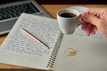 Hand reaching for a coffee cup on a wooden desk with a handwritten journal and laptop, a concept for creative work or study.