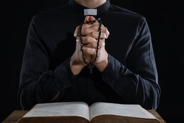 Priest with rosary and Holy Bible praying at table on black background, closeup