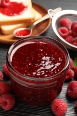 Sweet raspberry jam in glass jar, berries and bread slices on dark gray wooden table, closeup