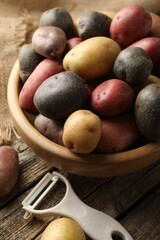 Different types of potatoes in bowl and peeler on wooden table, closeup