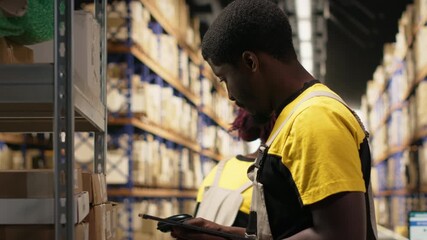 African American male employee scanning packages in a large warehouse, registering airway bills shipment details within industrial depot records. Using scanner for order fulfillment. Camera B.