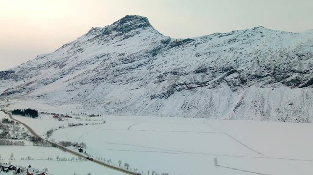 Aerial footage capturing the serene winter landscape of snow covered mountains, a winding road, and a vast frozen lake or field, reminiscent of the scenic beauty near Geiranger, Norway.