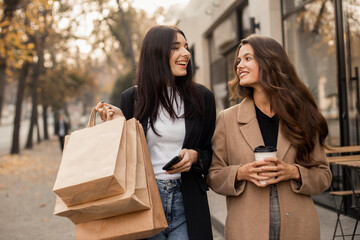 Two smiling women friends dressed in beige coat and jacket drinking coffee and using smartphone or phone, holding paper bags with purchases in hand after shopping walking along streets of autumn city.
