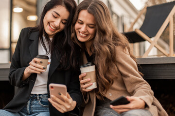 Two young women friends dressed in beige coat and jacket drinking coffee, having fun and chatting together and using smartphone sitting against the background of cafe in autumn seasons.