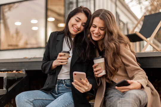 Two young women friends dressed in beige coat and jacket drinking coffee, having fun and chatting together and using smartphone sitting against the background of cafe in autumn seasons.