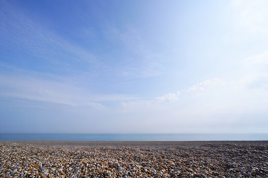 A clear blue sky with gentle clouds stretches above a vast calm ocean meeting a textured pebble beach.