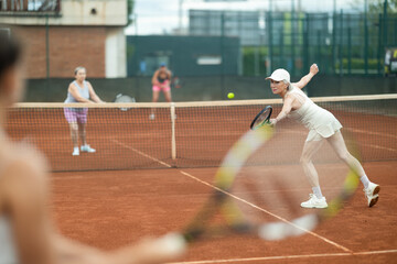 Rear view of emotional aged woman enthusiastically playing tennis outdoors in summer, preparing to hit forehand to return ball to opponent court during doubles match with female partner..