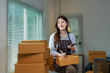 Asian female seller writing address on cardboard box preparing online delivery parcel, startup...