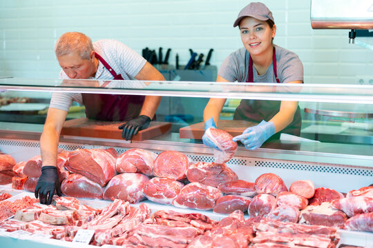 Adult man and young woman butchers in aprons put raw meat beef in butcher shop window