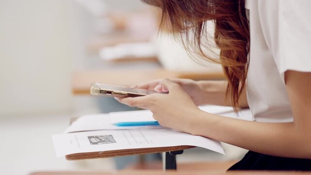 Asian female student using smartphone during classroom learning with paper document and pen on desk, representing tech-integrated education.