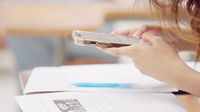 Female student using smartphone while study materials, documents, and pen are placed on school desk during class or self-study.