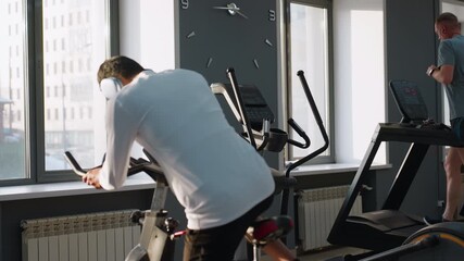 Young student in white sportswear walks to stationary bike and climbs on to begin cycling under sunlight streaming through gym windows, while background shows equipment and blurred workout scene - Powered by Adobe