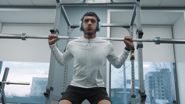 gym lover holding heavy barbell across shoulder while performing squat inside gym with bright window showing passing car and residential building in background