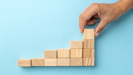 Hand stacking wooden blocks in ascending staircase pattern against light blue background business growth