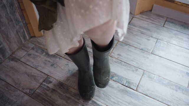 Elegant woman removing work boots while standing on rustic tiled floor, ruffled dress hem lifting to reveal bare calves, hands gripping boot edges, wood door frame and frost specks blurred
