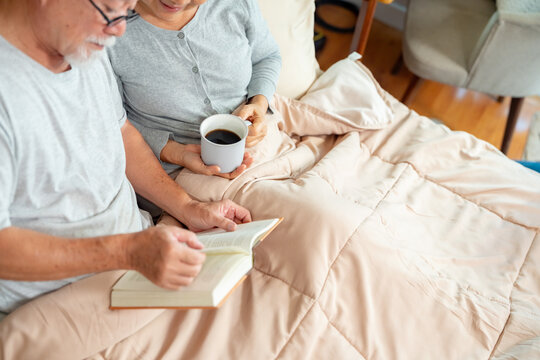 Happy Asian family senior couple relaxing and reading a book together on the bed in bedroom. Elderly husband and wife taking care each other. Retired people healthcare and mental health concept.