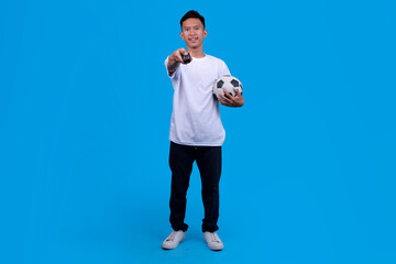 Happy Soccer Fan Holding Ball And Points TV Remote Ready To Watch Game On Blue Background