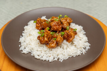 Close-up photograph of a plate of orange chicken on a serving of rice on a round brown plate on a round wooden board.