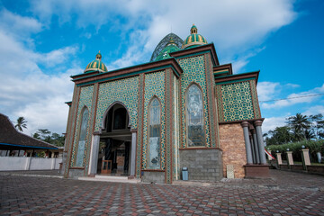 Beautiful Islamic mosque exterior featuring intricate gold geometric patterns on green walls, grand arched entrance, and multiple green domes under a blue sky.