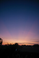 Beautiful twilight sky with vibrant gradient colors from deep blue to orange, featuring a delicate crescent moon above silhouetted trees and rooftops at dawn or dusk.