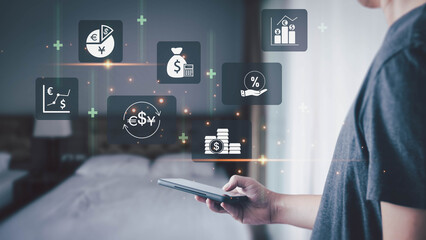 A man uses a smartphone to manage finances and investments. Holographic icons for currency exchange, stock charts, and money float above the device, illustrating modern financial technology at home.
