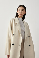 Woman in a cream oversized coat and striped top, standing in a minimal studio setting.