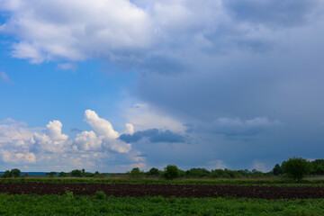 A rural landscape under a dynamic sky where bright blue skies with fluffy clouds meet a massive, dark gray rain cloud. In the foreground, a green field and freshly plowed soil are visible.