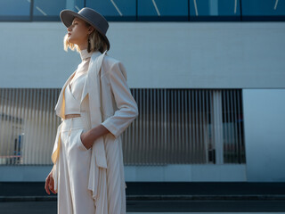 Woman in white outfit with hat standing outdoors by modern building.