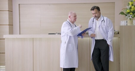 two caucasian male doctors consulting at hospital front desk while reviewing patient data on clipboard during slowmotion scene of collaboration and healthcare decision-making process - Powered by Adobe