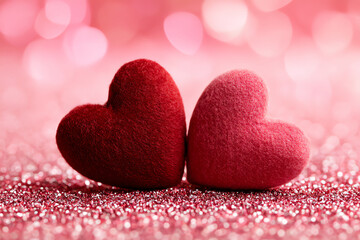Fototapeta premium Two soft, fuzzy hearts in red and pink sit side by side on a sparkling pink surface with a blurred pink bokeh background