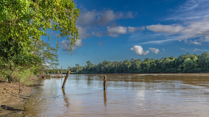 A tropical riverbed. There are rainforest thickets on the clay shores. There is a wooden boat pier above the water. The flag of Malaysia. The motorboat is moored. Blue sky, clouds. Kinabatangan 
River