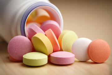 Colorful tablets spilling out of a white medicine bottle onto a wooden surface, showing various round pills in shades of pink, yellow, orange, and white