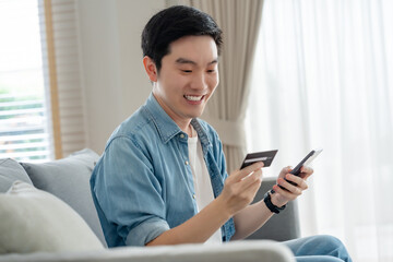 Young asian man sitting on sofa in living room, makes online banking payments through the internet from bank card on cell phone. Shopping online on mobile with credit card
